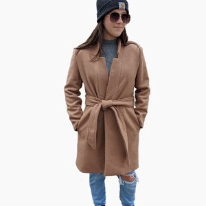 May include: A camel-colored wrap coat with a belt, worn with blue jeans and a black beanie.