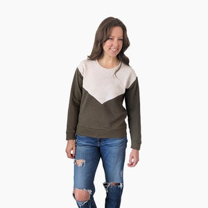 May include: A woman wearing a green and beige color block sweatshirt with long sleeves and a crew neck. She is wearing blue jeans with rips at the knees and brown ankle boots.