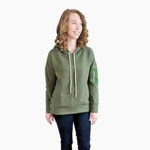 Puede incluir: Una sudadera con capucha verde con un bolsillo con cremallera en la manga izquierda. La sudadera tiene un cordón blanco y un corte holgado.