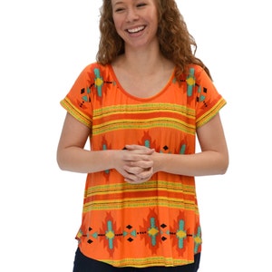 May include: A woman wearing a short-sleeved orange top with a geometric pattern in shades of turquoise, yellow, and black. The top has a scoop neck and a relaxed fit.