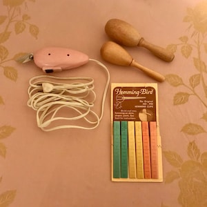 May include: A pink electric fabric cutter with a white cord, two wooden maracas, and a package of Hemming-Bird No-Pin Hemming Clips. The package has a brown background with yellow text and shows six colorful clips.