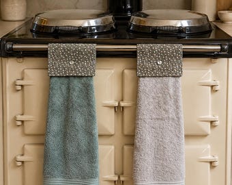 May include: Two hanging kitchen towels draped over a stove handle. One towel is sage green and the other is light gray. Both feature a top fabric panel with a dark gray and white polka dot pattern, secured by a single white button on each.