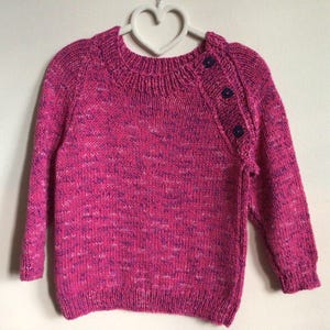 May include: A knitted pink sweater with a crew neck and long sleeves. The sweater has a buttoned shoulder detail with three dark purple buttons. The yarn has a speckled texture with shades of pink, purple, and white.