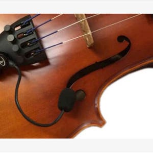 May include: A close-up of a violin with a black pickup attached to the bridge. The pickup has a wire that connects to a small microphone.