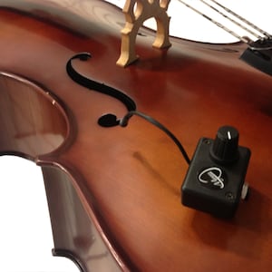 May include: A black cello pickup with a volume knob attached to a brown cello. The pickup is connected to the cello with a black wire.