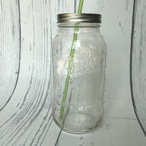 May include: A clear glass mason jar with a silver lid and a green straw. The jar has the embossed word "Ball" on the front. The jar is empty and sits on a white and gray wooden surface.