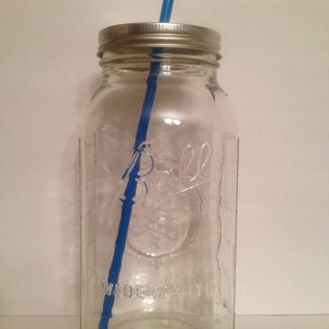 May include: A clear glass mason jar with a silver lid and a blue straw. The jar has the word "Ball" and "Wide Mouth" embossed on the front. The jar is empty and ready to be filled with a beverage.