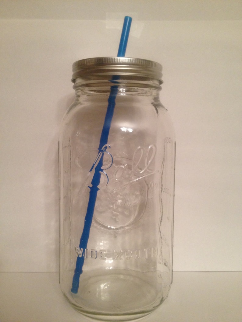 May include: A clear glass mason jar with a silver lid and a blue straw. The jar has the word "Ball" and "Wide Mouth" embossed on the front. The jar is empty and ready to be filled with a beverage.