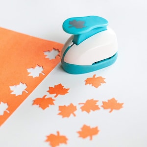 May include: A blue and white leaf punch with a gray leaf design on the handle. The punch is surrounded by orange paper leaves that have been punched out.