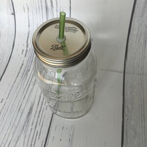 May include: A clear glass Ball jar with a silver lid and a green straw. The lid has the words "craft seal" and "for quality freshness" printed on it. The jar is sitting on a white wooden surface.
