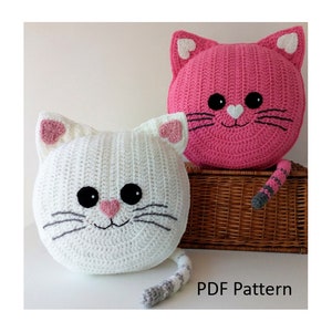 May include: Two crocheted cat pillows, one white and one pink, with black eyes, gray whiskers, and pink hearts on their ears. The pillows are sitting on a wicker basket and have gray and white tails.