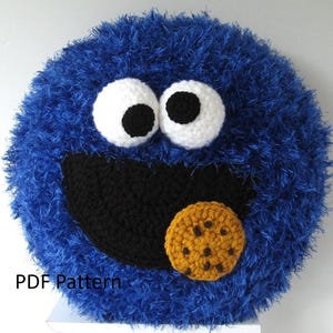 May include: Blue crocheted plush toy shaped like Cookie Monster from Sesame Street. The toy has large white eyes with black pupils, a black mouth, and a brown chocolate chip cookie in its mouth. The toy is made with a fuzzy blue yarn.