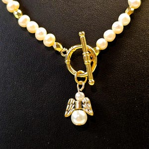 May include: A pearl necklace with a gold clasp and a gold angel pendant. The necklace features cream-colored pearls and gold accents. The angel pendant has a pearl body and gold wings.