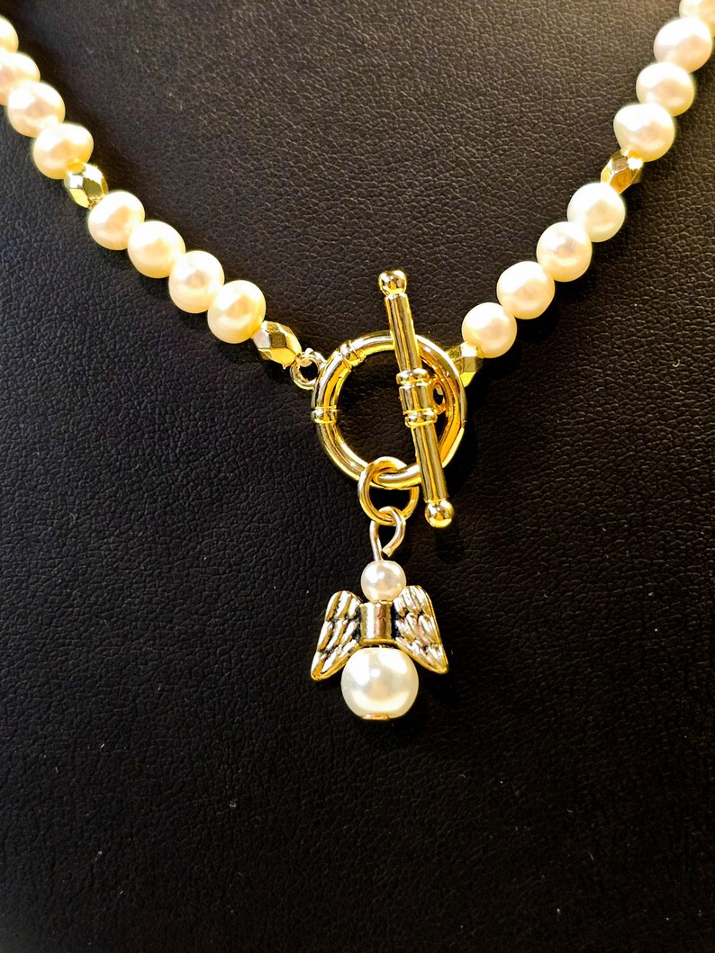 May include: A pearl necklace with a gold clasp and a gold angel pendant. The necklace features cream-colored pearls and gold accents. The angel pendant has a pearl body and gold wings.