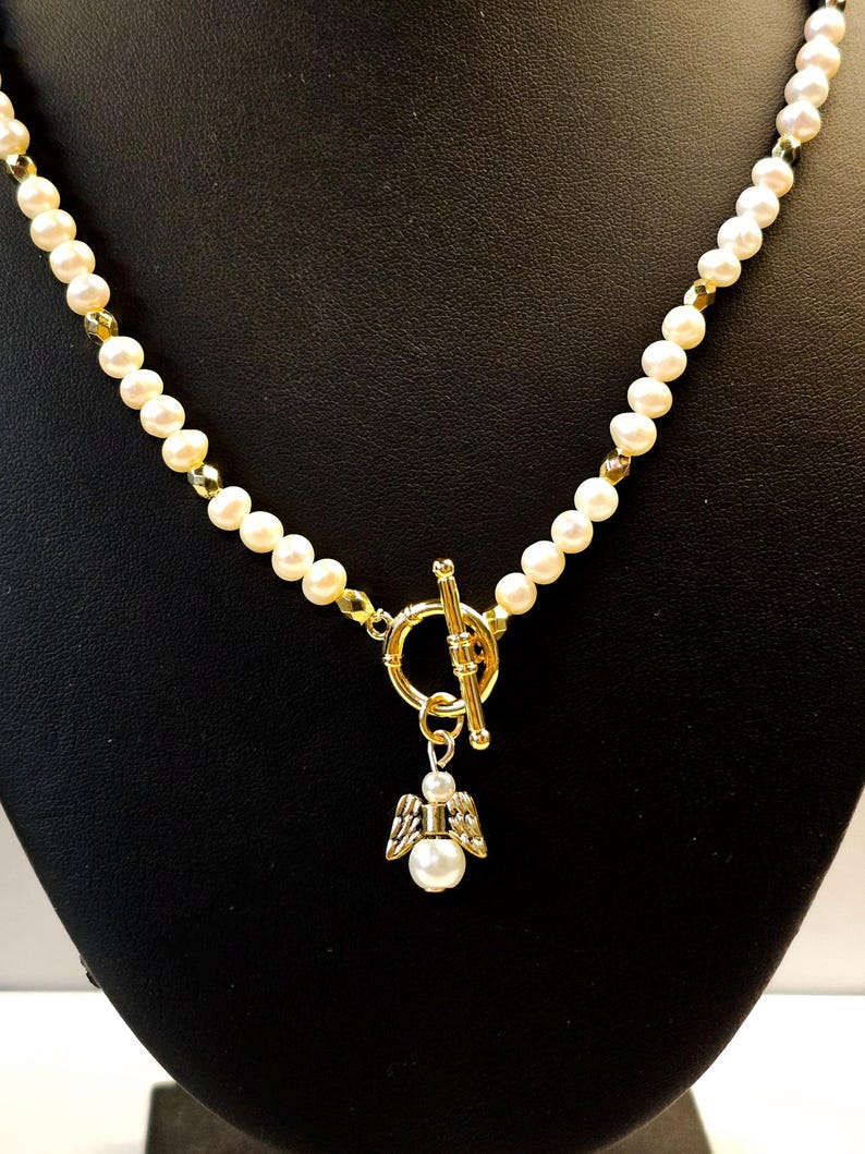 May include: A pearl necklace with a gold clasp and a small angel pendant. The necklace features cream-colored pearls and gold-colored accents. The angel pendant has a pearl body and gold-colored wings.