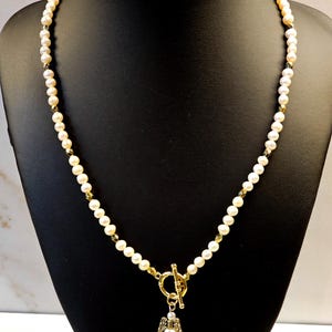 May include: A delicate pearl necklace with a gold-colored toggle clasp and a small angel pendant. The necklace features small, round pearls and gold-colored accents. The angel pendant has a pearl body and gold-colored wings.