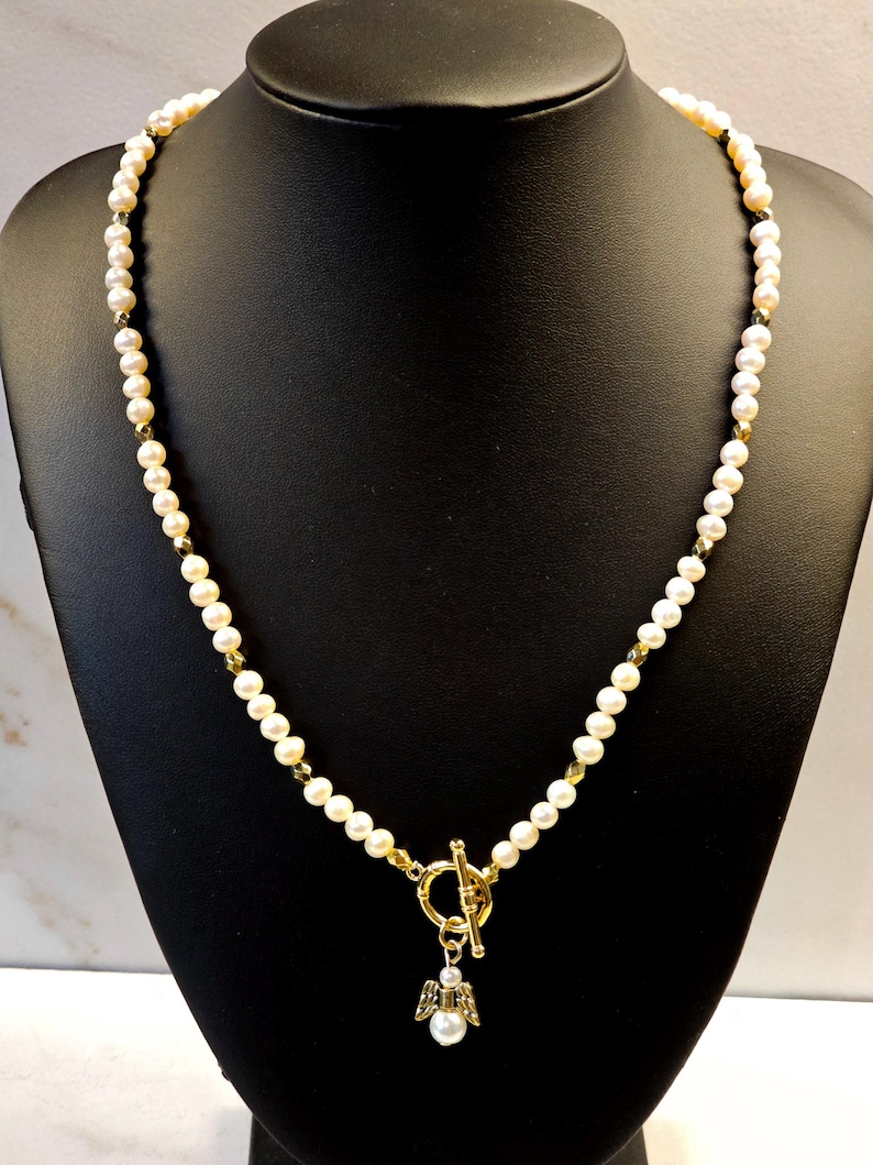 May include: A delicate pearl necklace with a gold-colored toggle clasp and a small angel pendant. The necklace features small, round pearls and gold-colored accents. The angel pendant has a pearl body and gold-colored wings.