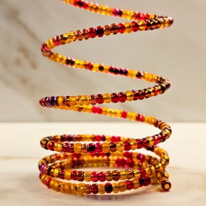 Peut inclure: Un bracelet en spirale composé de petites perles colorées. Les perles sont un mélange de rouge, d'orange et de jaune, créant un aspect chaleureux et vibrant. Le bracelet est enroulé en spirale, avec plusieurs boucles.