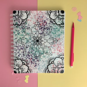 May include: A white spiral bound notebook with a colourful mandala design. The cover features a large mandala in the centre with smaller mandalas around the edges. The background is white with splashes of pink, blue, and green.