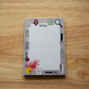 May include: A grey notepad with a white interior and a border of various illustrations, including a magnifying glass, a paintbrush, a DNA strand, a footprint, a scalpel, and a blood splatter. The notepad has the word "Notes" printed at the top.