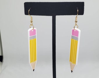 May include: A pair of gold dangle earrings with a white pencil charm. The pencil charm has a pink eraser, gray band, and yellow body with lines.