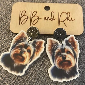 May include: A pair of earrings featuring a Yorkshire Terrier dog design. The earrings have a black glitter top and are attached to a wooden tag that reads "BB and Rhi". The dog's fur is black, brown, and tan.