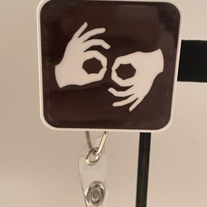 May include: A white square badge with a black background featuring a sign language symbol for the letter "I" in white. The badge is attached to a clear plastic retractable badge holder with a silver clip.