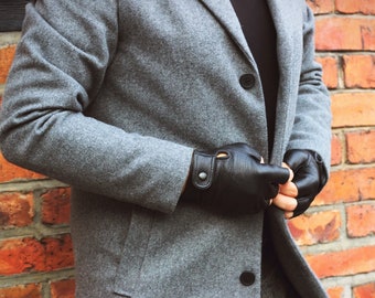 Puede incluir: Un abrigo de lana gris con un guante de cuero negro en la mano izquierda y un guante de cuero negro en la mano derecha. La persona lleva una camisa negra debajo del abrigo.