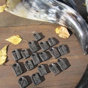 Puede incluir: Una colección de 20 colgantes de runas negras con diferentes símbolos tallados en ellos. Los colgantes están dispuestos sobre una superficie de madera con un par de cuernos en el fondo.