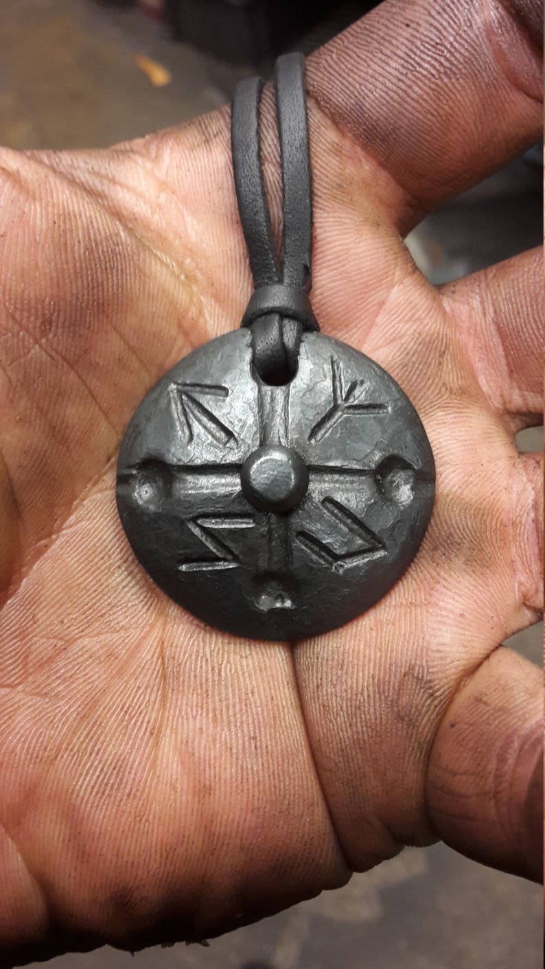 K&ouml;nnte beinhalten: Eine Halskette aus schwarzem Lederband mit einem runden, schwarzen Metallanh&auml;nger mit einem geschnitzten Design. Der Anh&auml;nger hat eine erhabene Mitte und eine strukturierte Oberfl&auml;che.