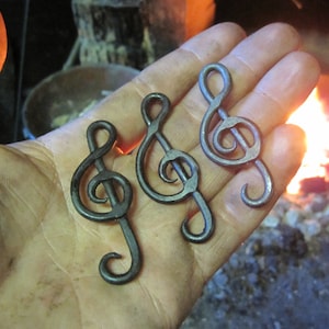 May include: Three metal treble clef music note charms, hand-forged and polished, with a rustic finish. The charms are held in a hand against a blurred background of a glowing fire.