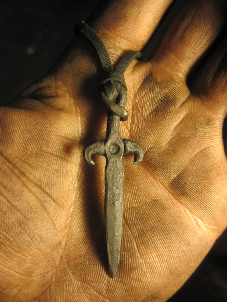 May include: A silver sword pendant with a black leather cord. The sword has a pointed tip and a cross-shaped guard.