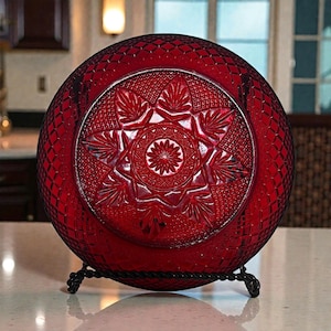 May include: A ruby red glass plate with intricate star and leaf patterns. The plate is displayed on a black metal stand. The plate's design features a central starburst surrounded by detailed leaf and diamond patterns.