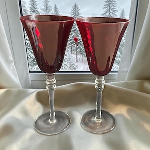 May include: A pair of wine glasses featuring deep red, iridescent bowls with a subtle rippled texture. Each glass has a clear, slender stem with a decorative ring detail near the top and a wide, circular clear glass base for stability.