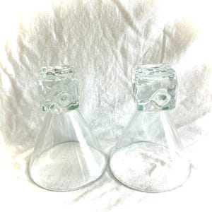 May include: Two clear glass hourglasses with square tops and conical bases. The hourglasses are filled with white sand. The background is a white, textured fabric.
