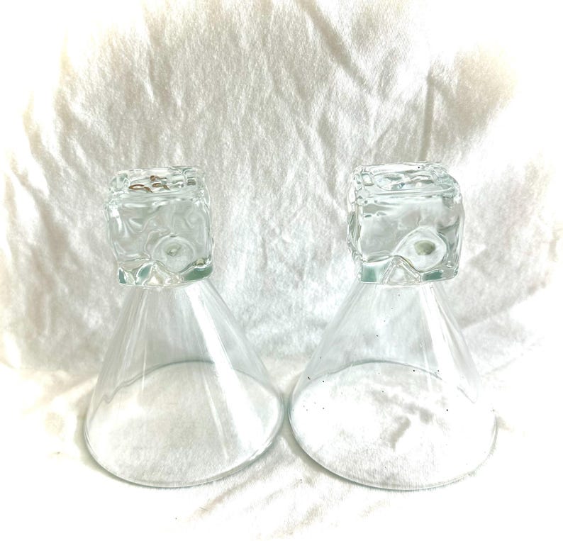 May include: Two clear glass hourglasses with square tops and conical bases. The hourglasses are filled with white sand. The background is a white, textured fabric.