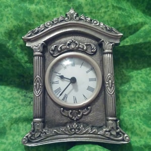 Op de afbeelding: Een zilverkleurige, sierlijke mantelklok met een witte wijzerplaat en zwarte Romeinse cijfers. De klok heeft decoratieve zuilen en krullen, en de wijzers wijzen naar ongeveer 4:15 uur. De klok staat tegen een groene achtergrond.