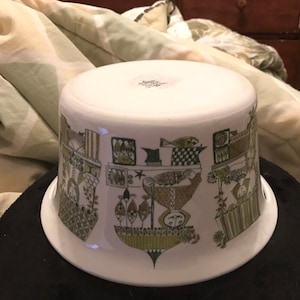 Puede incluir: Un bol de servir de cerámica blanca con un patrón geométrico verde y marrón que presenta peces y pájaros estilizados. El bol tiene un borde ancho y una base redondeada.