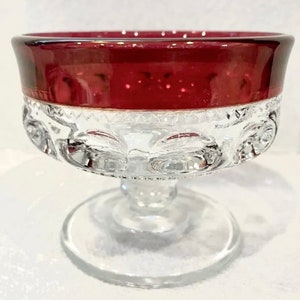 May include: A clear glass dessert dish with a ruby red rim. The bowl has a decorative, patterned design around the middle and sits on a clear, round base. The dish is suitable for serving desserts.