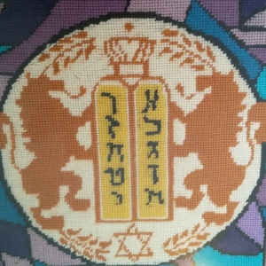 May include: A framed embroidery featuring a white circle with two brown lions flanking two yellow scrolls with Hebrew text. The circle is surrounded by a wreath of green leaves and a crown at the top. The background is a colorful mosaic of purple, blue, and green.
