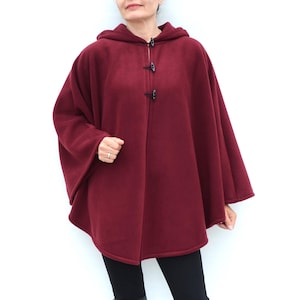 May include: A burgundy hooded cape with three dark toggle closures down the front. The cape is made of a soft material and has a relaxed fit. The person is wearing black leggings and black knee-high boots.