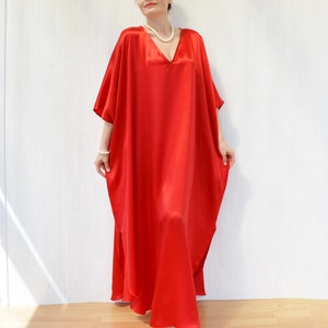 May include: A flowing, floor-length red satin caftan dress with a V-neckline and short sleeves. The dress has a loose, comfortable fit and a split on the side. A pearl necklace and bracelet are also visible.