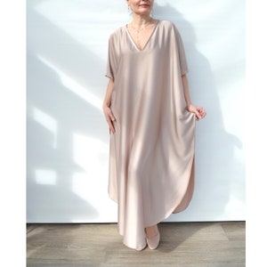 May include: A long, flowing, beige maxi dress with a V-neck and short sleeves. The dress has a relaxed fit and a slightly asymmetrical hemline.