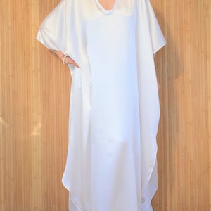 May include: A white, flowing kaftan dress with a V-neckline and short sleeves. The dress is made of a silky fabric and has a relaxed fit.