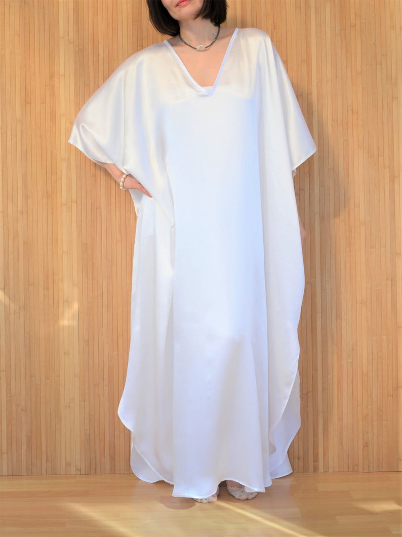 May include: A white, flowing kaftan dress with a V-neckline and short sleeves. The dress is made of a silky fabric and has a relaxed fit.