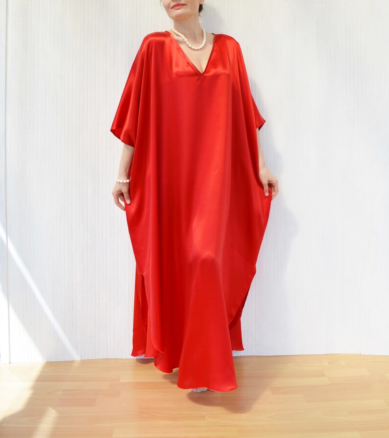 May include: A flowing, floor-length red satin caftan dress with a V-neckline and short sleeves. The dress has a loose, comfortable fit and a split on the side. A pearl necklace and bracelet are also visible.