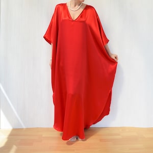 May include: A vibrant red, flowing caftan dress with a V-neckline. The dress is made of a shiny, silky material and has wide sleeves. The model is wearing a pearl necklace. The dress is floor-length.