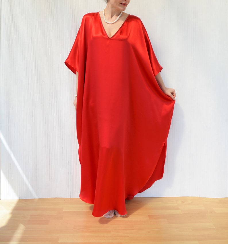 May include: A vibrant red, flowing caftan dress with a V-neckline. The dress is made of a shiny, silky material and has wide sleeves. The model is wearing a pearl necklace. The dress is floor-length.