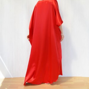 May include: A flowing, vibrant red caftan dress. The dress features wide sleeves and a loose, comfortable fit. The fabric has a silky sheen, suggesting a luxurious material. The dress is floor-length, creating an elegant silhouette.