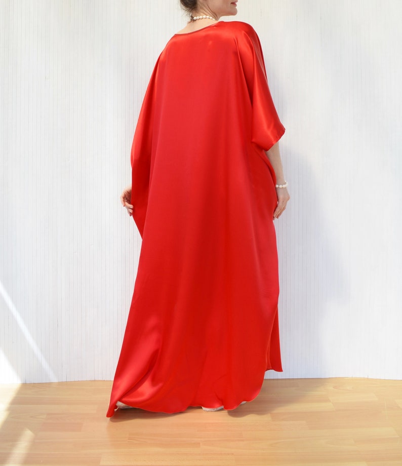 May include: A flowing, vibrant red caftan dress. The dress features wide sleeves and a loose, comfortable fit. The fabric has a silky sheen, suggesting a luxurious material. The dress is floor-length, creating an elegant silhouette.